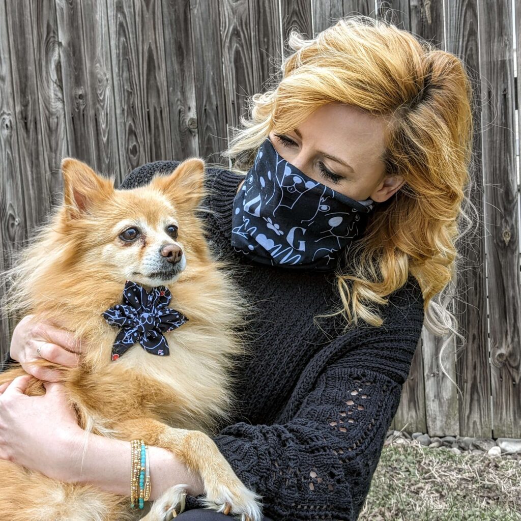 Blonde woman wearing a bandana over her face holds a dog wearing a Huxley & Kent bowtie