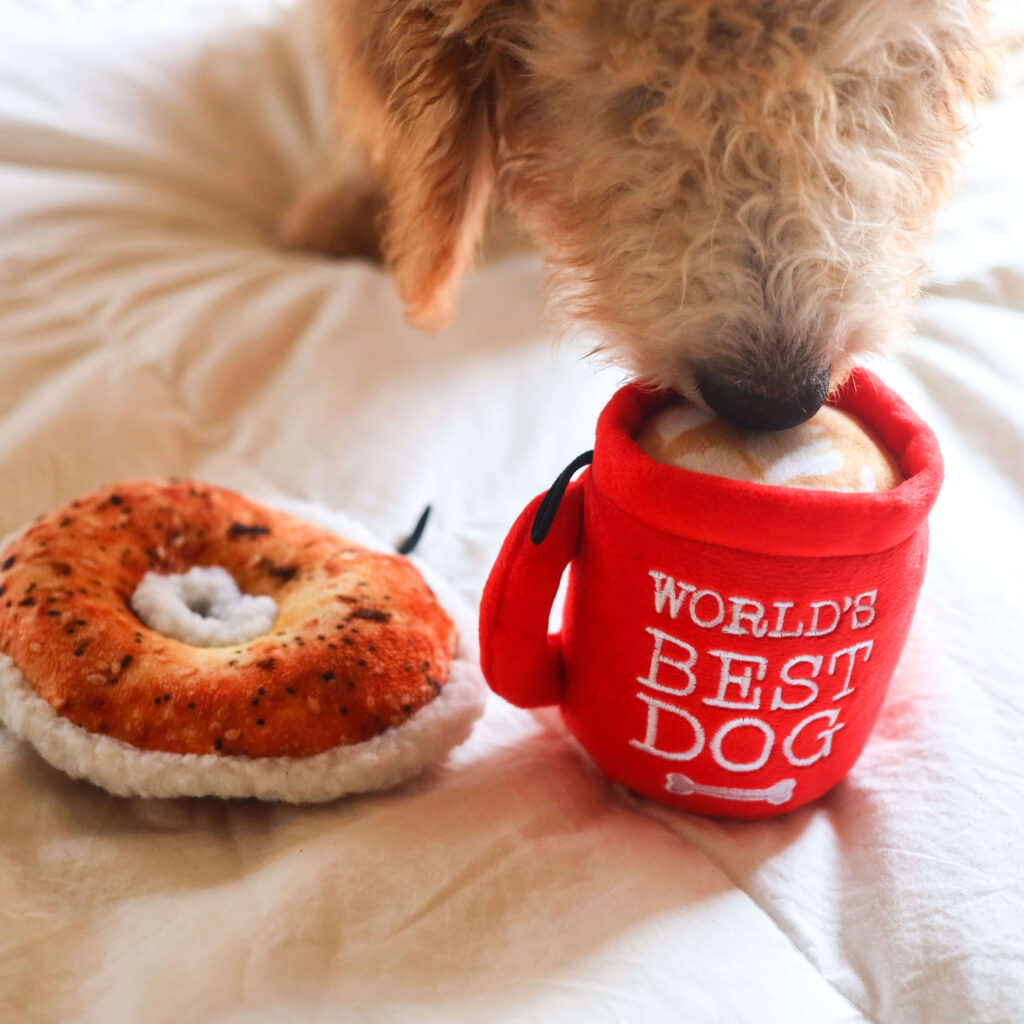 Golden doodle dog sniffs a Huxley & Kent dog toy shaped as a red mug, with a Huxley & Kent donut toy beside it