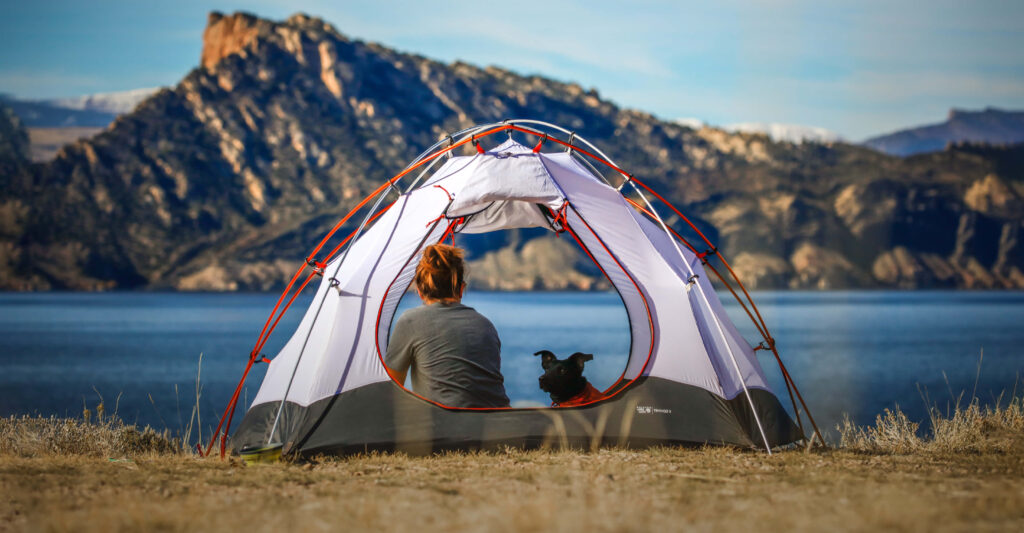 A woman and her dog sit in a tent beside a lake on a sunny day while on a pet-friendly camping vacation.