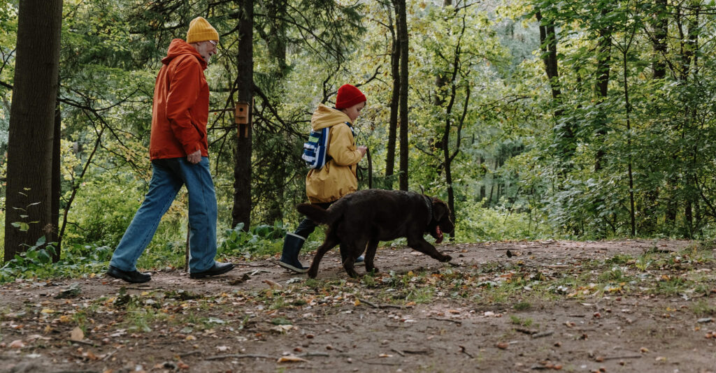 A parent and their child walk their black dog along a trail in a lush green forest.