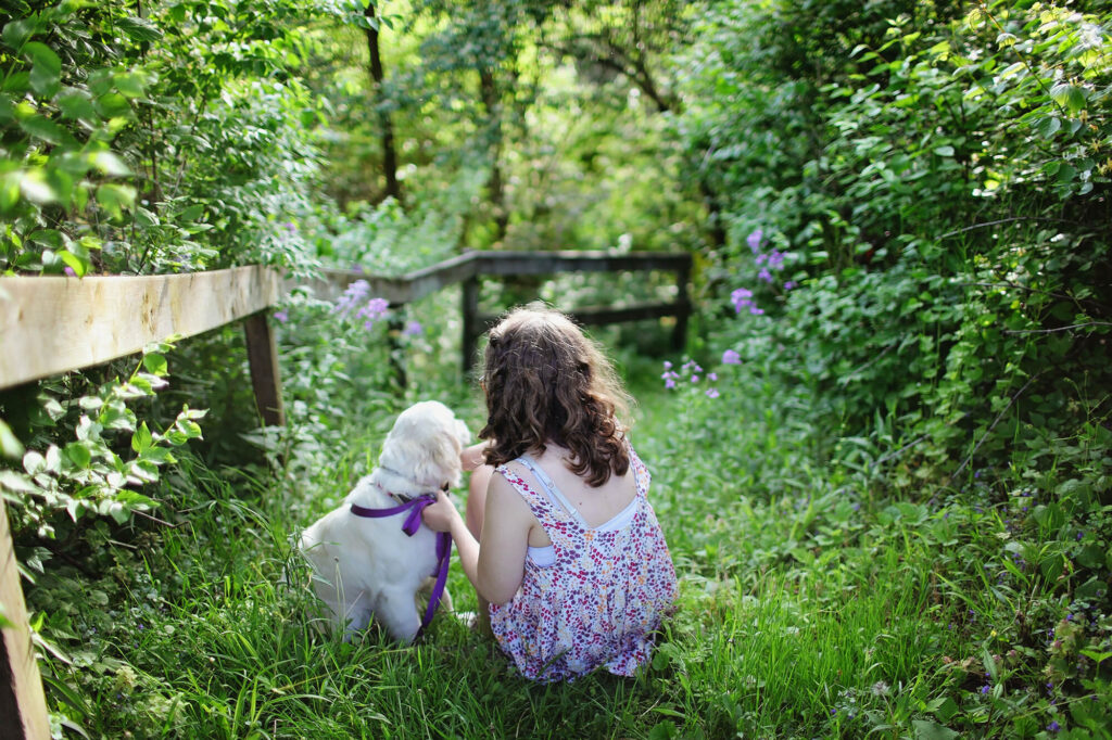 A young girl in a purple dress holds the purple colour of a golden retriever puppy as they look at a lush green garden during the Spring.