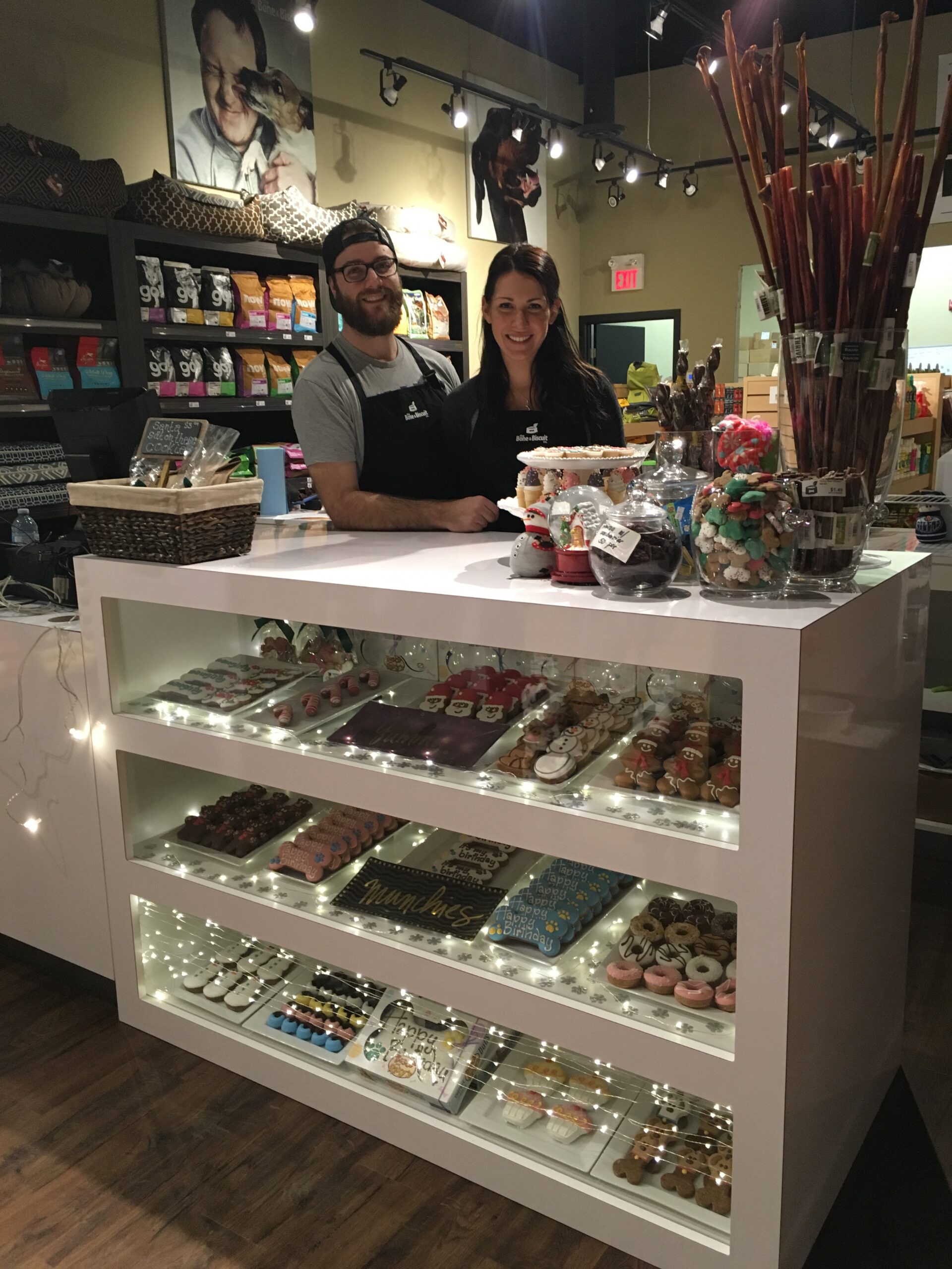 Amanda and Travis Sarvas stand behind the counter of their Bone & Biscuit Leduc location surrounded by dog bakery items and pet supplies.