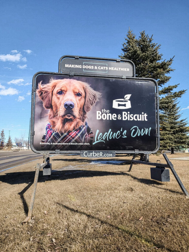 A large billboard with a golden retriever advertising for the Bone & Biscuit Leduc location stands on a grassy field by the side of the road with a pine tree in the background.