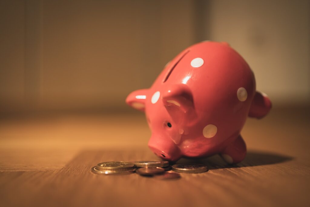 A pink piggy bank with white polka dots stands tilted on a wooden table with coins spilling out.