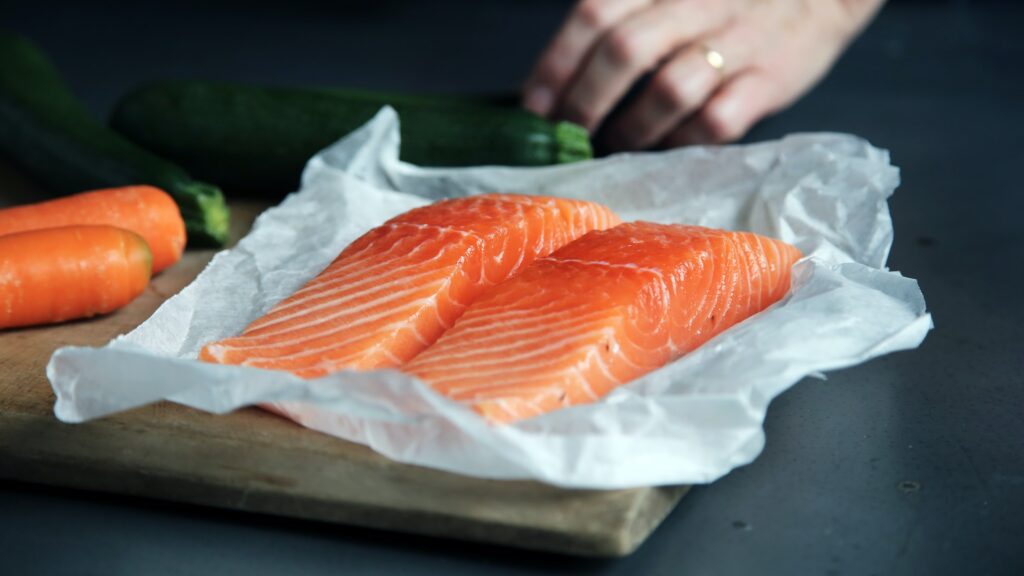 Two fillets of raw salmon on wax paper sits on a cutting board with carrots in the background for a raw food diet for Rawgust.