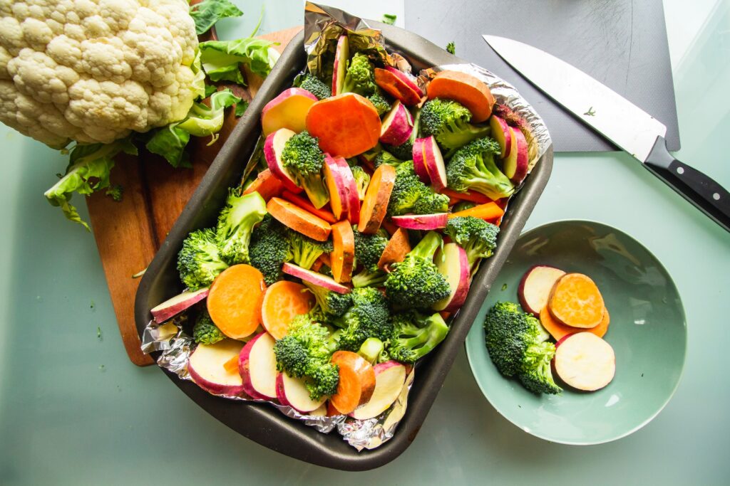 A baking dish loaded with raw veggies of broccoli, potatoes and yams sits on a cutting board, with another bowl of veggies beside it, a cauliflower head above, and a knife on the side.