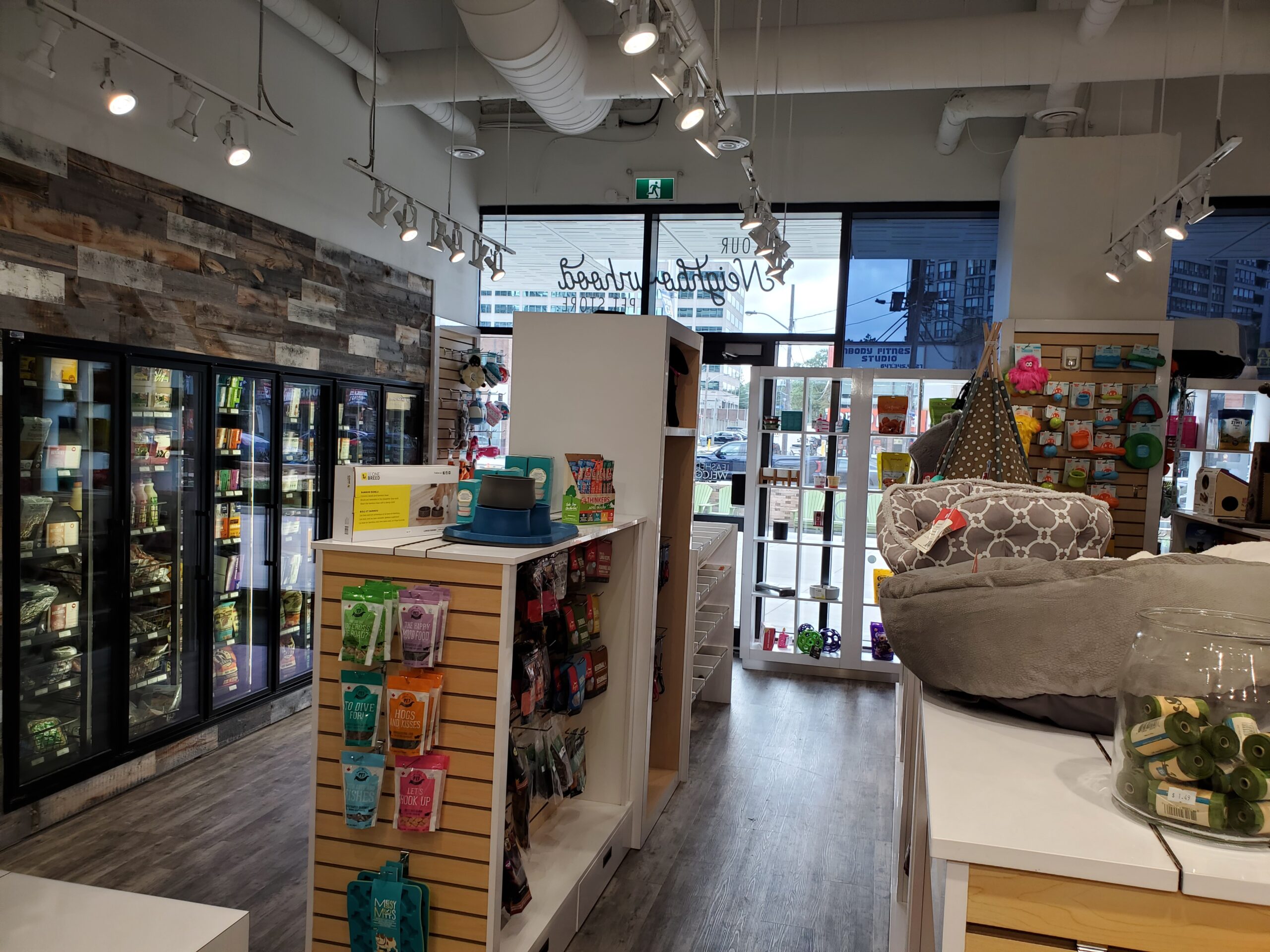 Inside the Bone & Biscuit Yonge Sheppard store, displaying it's wood-style floors and shelves of dog food, cat food, pet beds and pet treats in a brightly lit store.