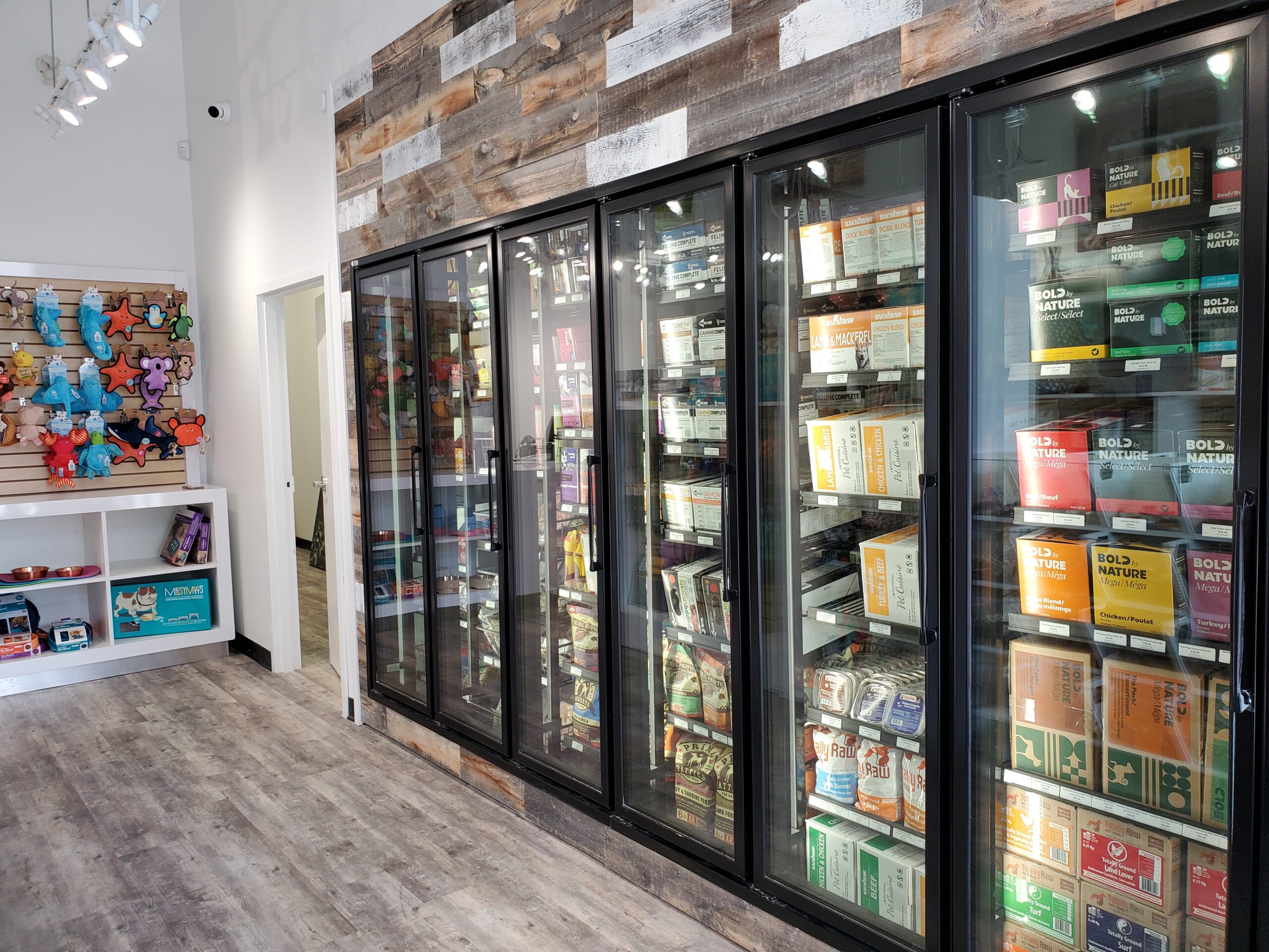 Inside the Bone & Biscuit Yonge Sheppard store, displaying it's wood-style floors and displaying its frozen and refrigerated storage area with organized shelves and clear glass, which are full of dog food and cat food in a brightly lit store.