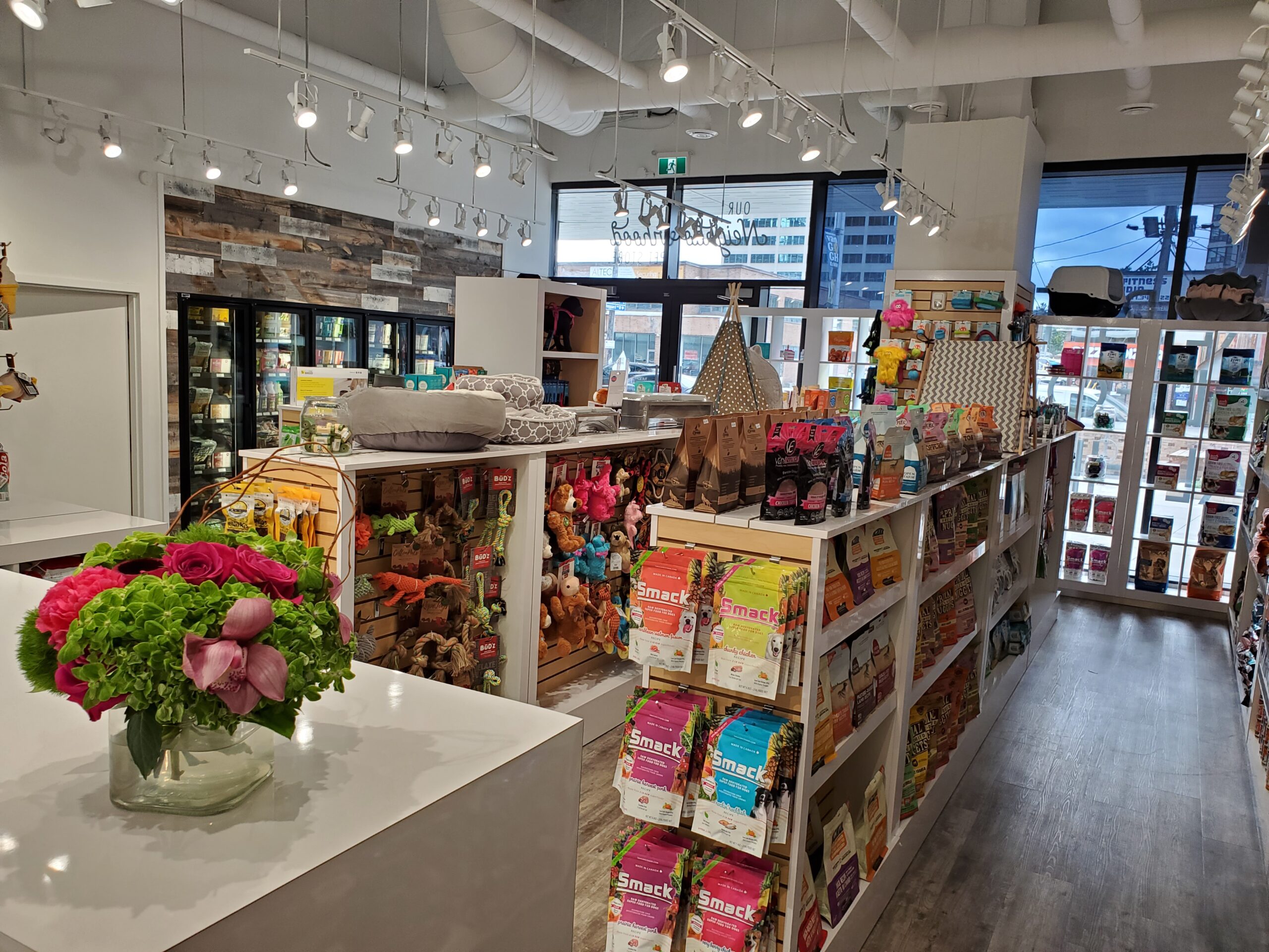 Inside the Bone & Biscuit Yonge Sheppard store, displaying it's wood-style floors and a bouquet of flowers on the counter and shelves of dog food, cat food, pet beds and pet treats in a brightly lit store.