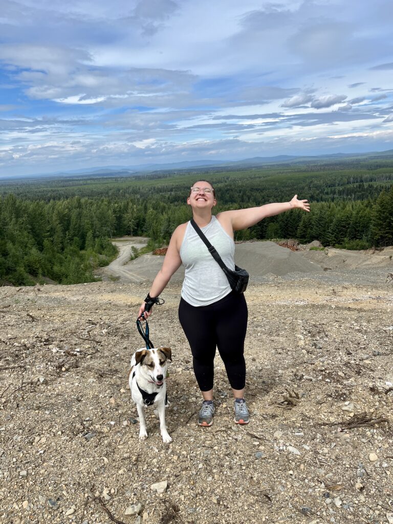 Courtney, the owner of the new Bone & Biscuit Garneau, stands smiling with her arms wide and holding a leash to her dog, Ellie, while hiking outdoors in Edmonton Alberta.
