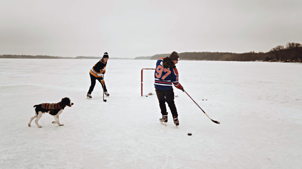 Two people in warm clothing and hockey jerseys, one wearing an Edmonton Oilers McDavid jersey, play ice hockey outdoors in Canada with their dog.