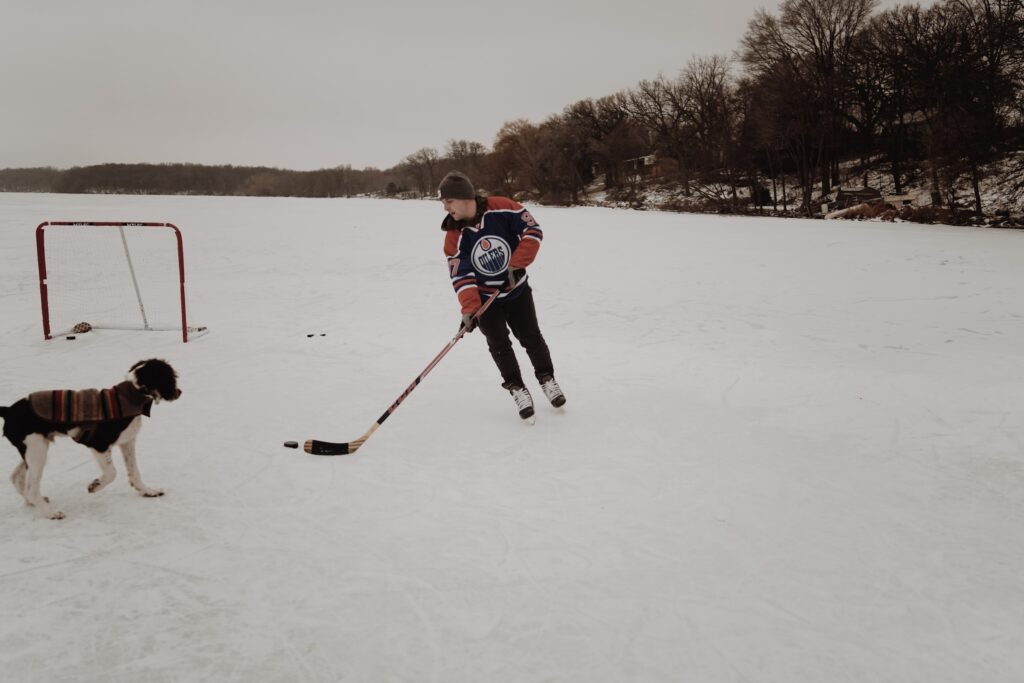 A man in a toque and Edmonton Oilers jersey plays outdoor ice hockey in Canada with his dog.