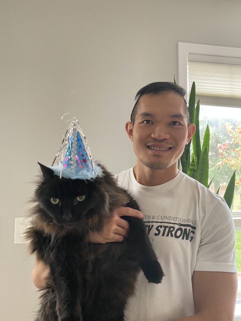 A photo of Phum, a black long-haired cat, wearing a birthday party hat while his owner, Fung, holds him and smiles for a birthday photo. Fung is a new store owner of the Bone & Biscuit Okotoks location.
