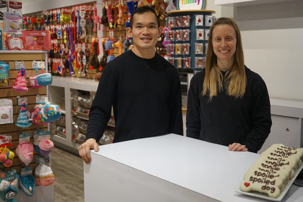 A photo of Bone & Biscuit Okotoks' new store owners, Amber & Fung, standing behind the store's front counter smiling in their Okotoks pet supply store.