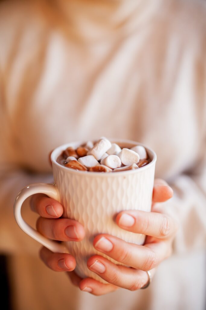 A woman in a white sweater holding a white mug with hot chocolate topped with marshmallows.