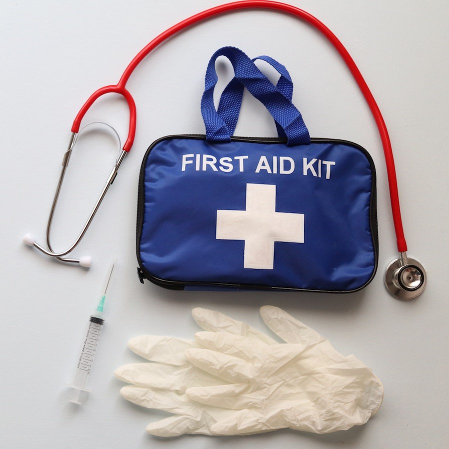 A first aid kit with a stethoscope, gloves and a syringe on a white background. Photo by Roger Brown: https://www.pexels.com/photo/first-aid-kit-on-white-background-5664736/
