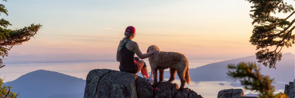 A woman in hiking gear sits on a rock with her golden retriever dog looking out at the sunset on a hike for their adventure day dog activity together.