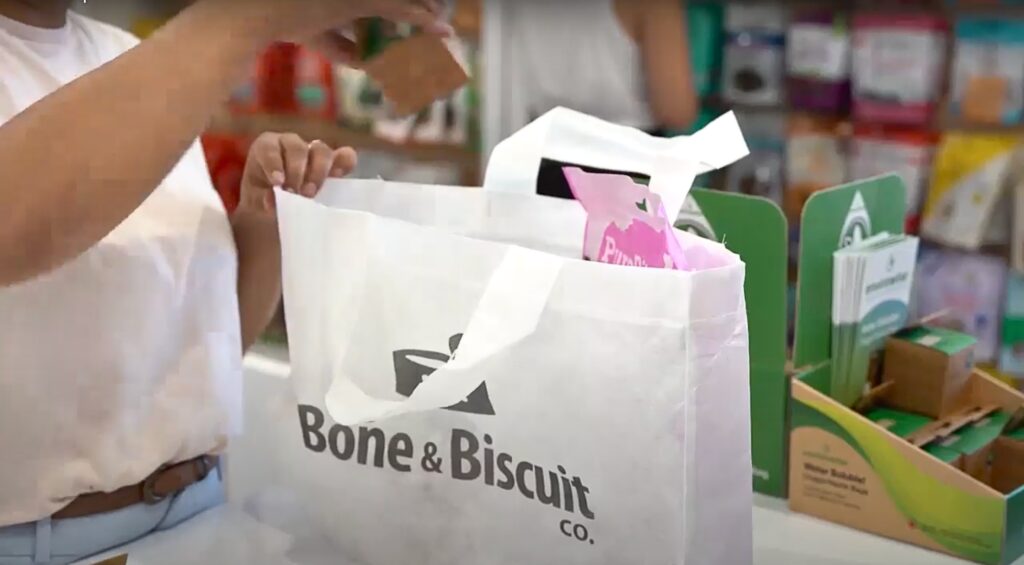 Akanksha, the owner of Bone & Biscuit Fraser Heights Surrey pet store, wears a white t-shirt and places a box of Envirowise Solubag biodegradable dog poop bags into their water soluble shopping bags with the Bone & Biscuit logo on the front.