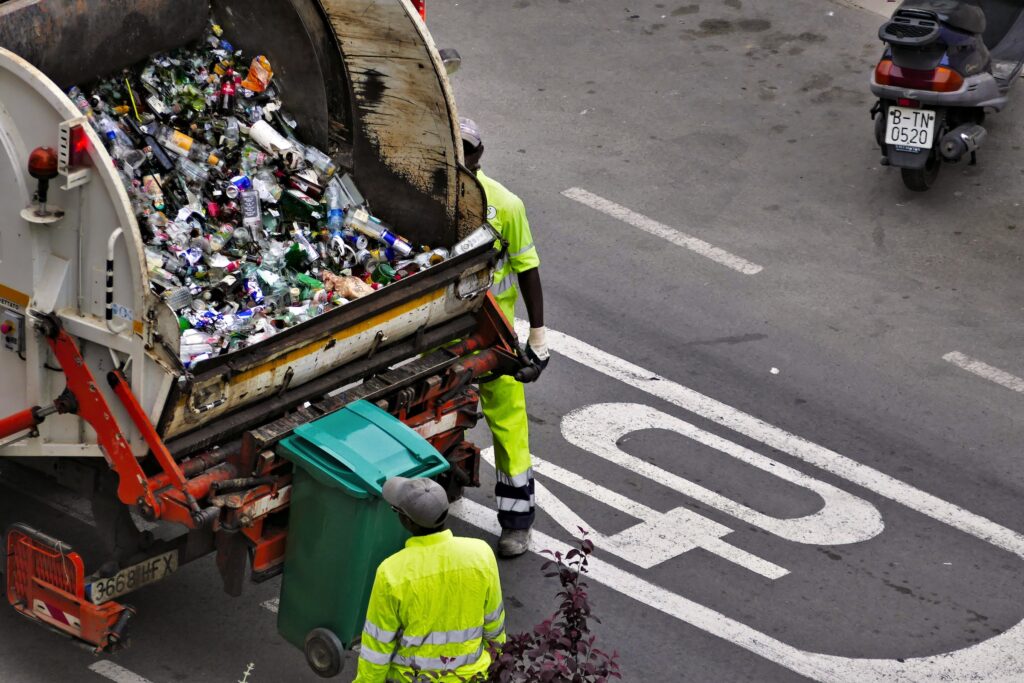 Two people in yellow safety uniforms load garbage bins into a garbage truck on the side of a street, getting garbage and waste like dog poop bags loaded to go to the landfill.