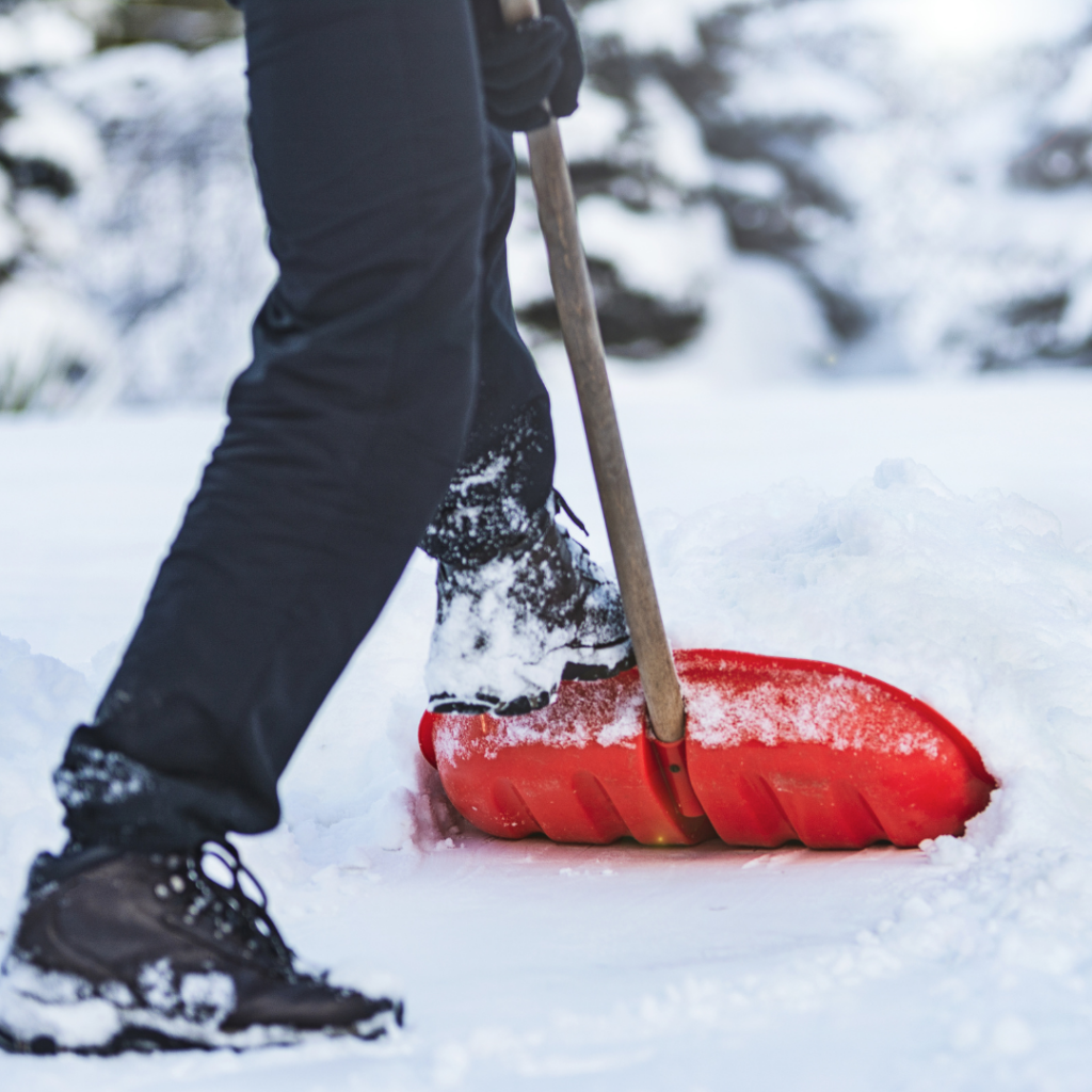 A person wearing black pants and boots uses a red snow shovel to clear a sidewalk from snow in winter.