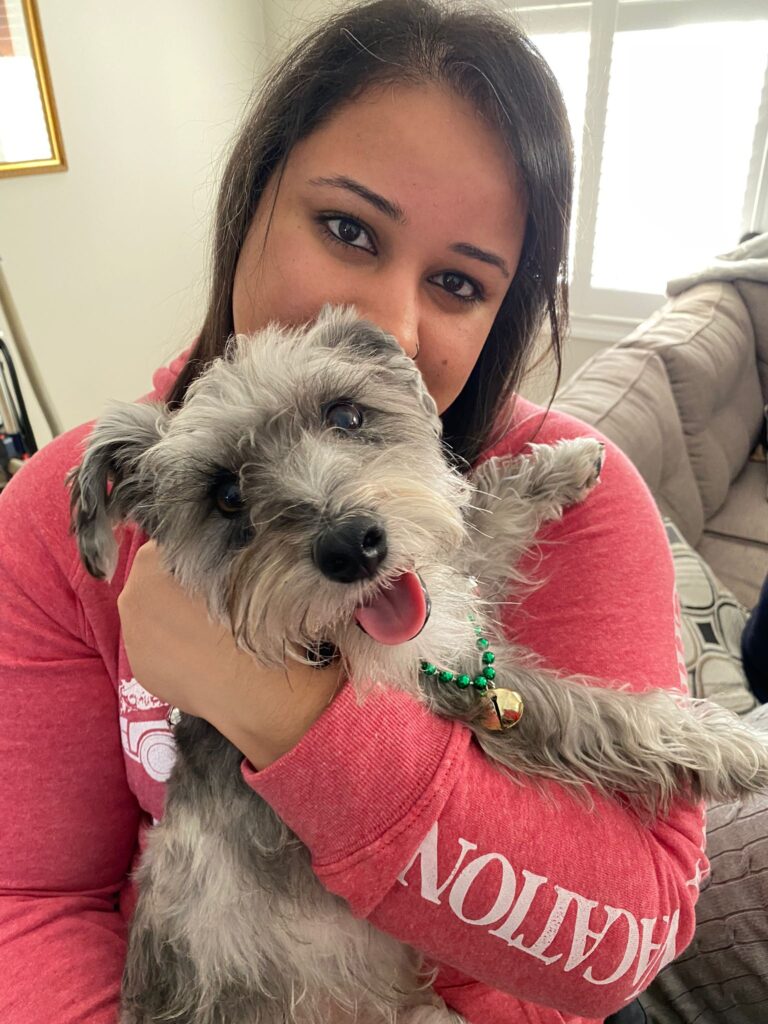 Jas, the store owner of Bone & Biscuit The Well in Toronto, sits on a couch in a reddish-pink shirt hugging her dog Dash.