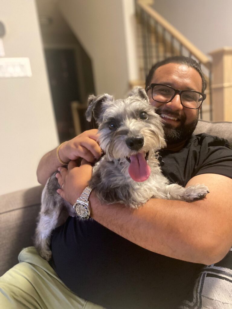Sunil, the owner of the Well Bone & Biscuit, sits on a chair in a black shirt while hugging his dog Dash and smiling for the camera.