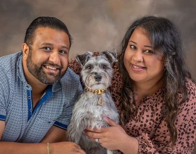 A photo of Jas and Sunil, the owners of the new Toronto pet store Bone & Biscuit The Well, posing for a photo and smiling with their schnauzer dog named Dash.