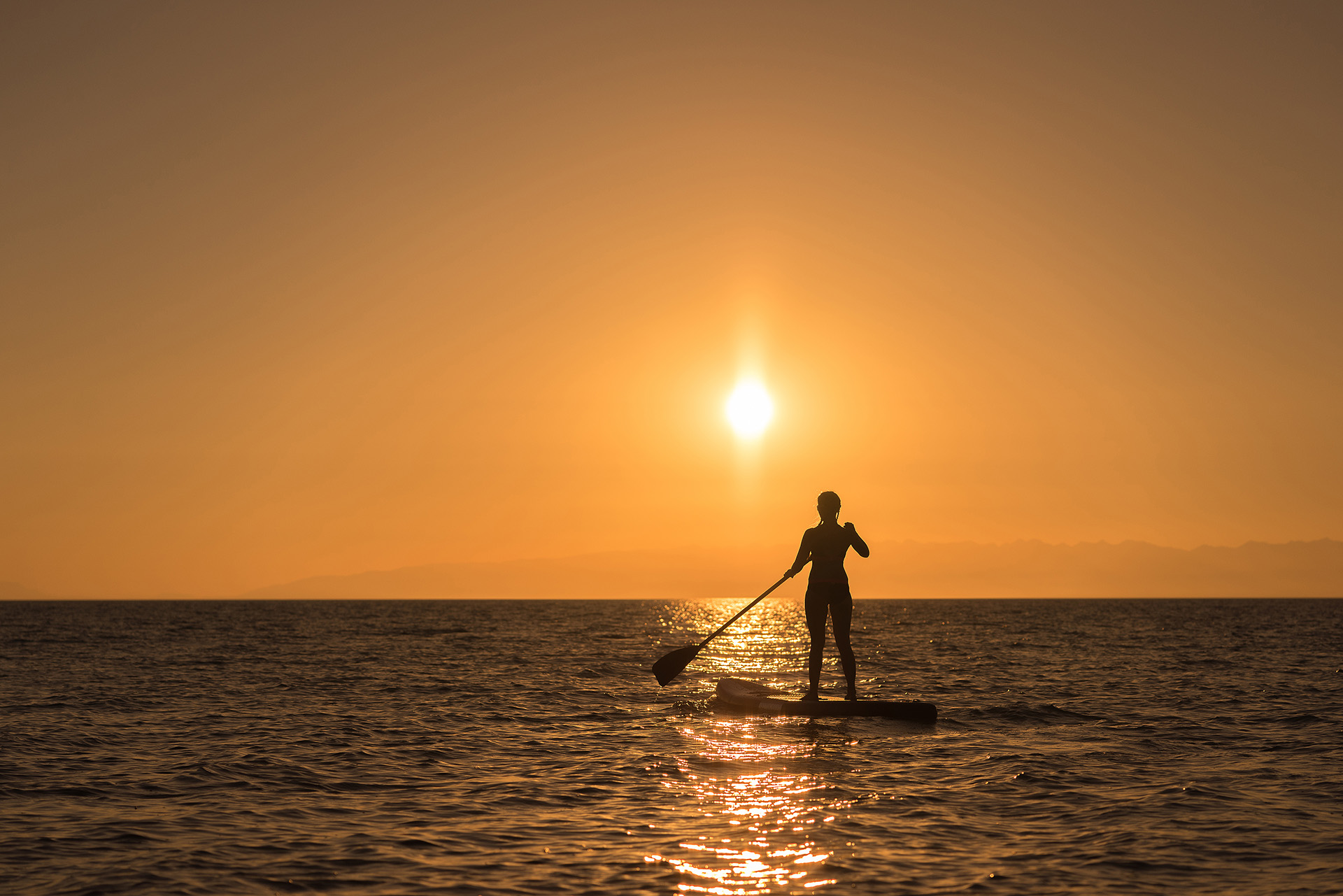 Person paddleboarding at sunset on calm water, silhouetted by an orange sky near Caerula Mar Club, South Andros, Bahamas.